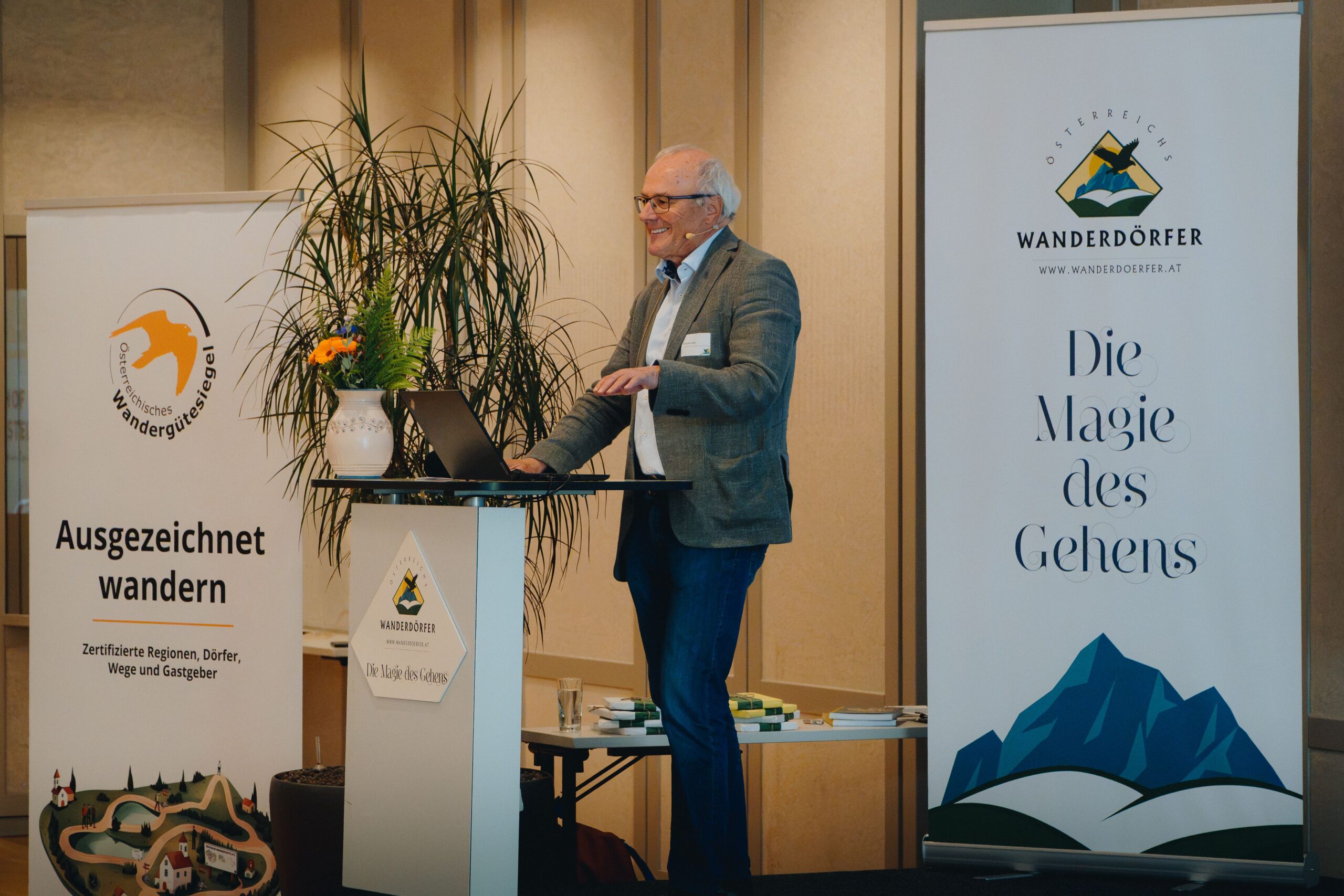 Ein Mann in einem Blazer hält eine Rede an einem Rednerpult, neben dem Banner mit Wandermotiven und eine Topfpflanze stehen.