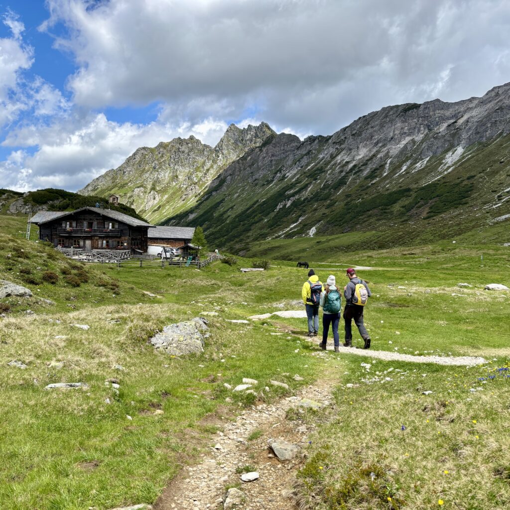 Drei Wanderer gehen auf einem Bergpfad auf eine Holzhütte zu, umgeben von grasbewachsenen Hügeln und felsigen Gipfeln.