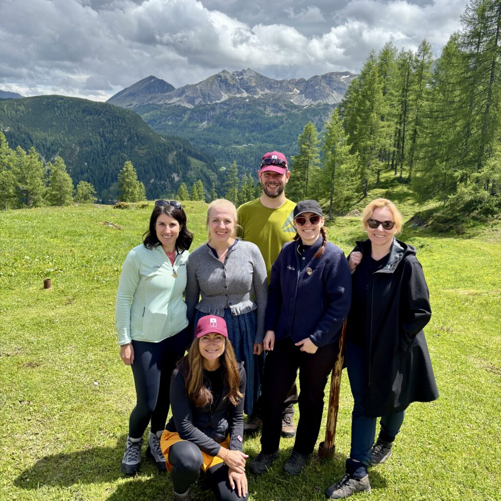 Sechs Menschen posieren und lächeln in einer bergigen Graslandschaft mit Bäumen und bewölktem Himmel im Hintergrund.