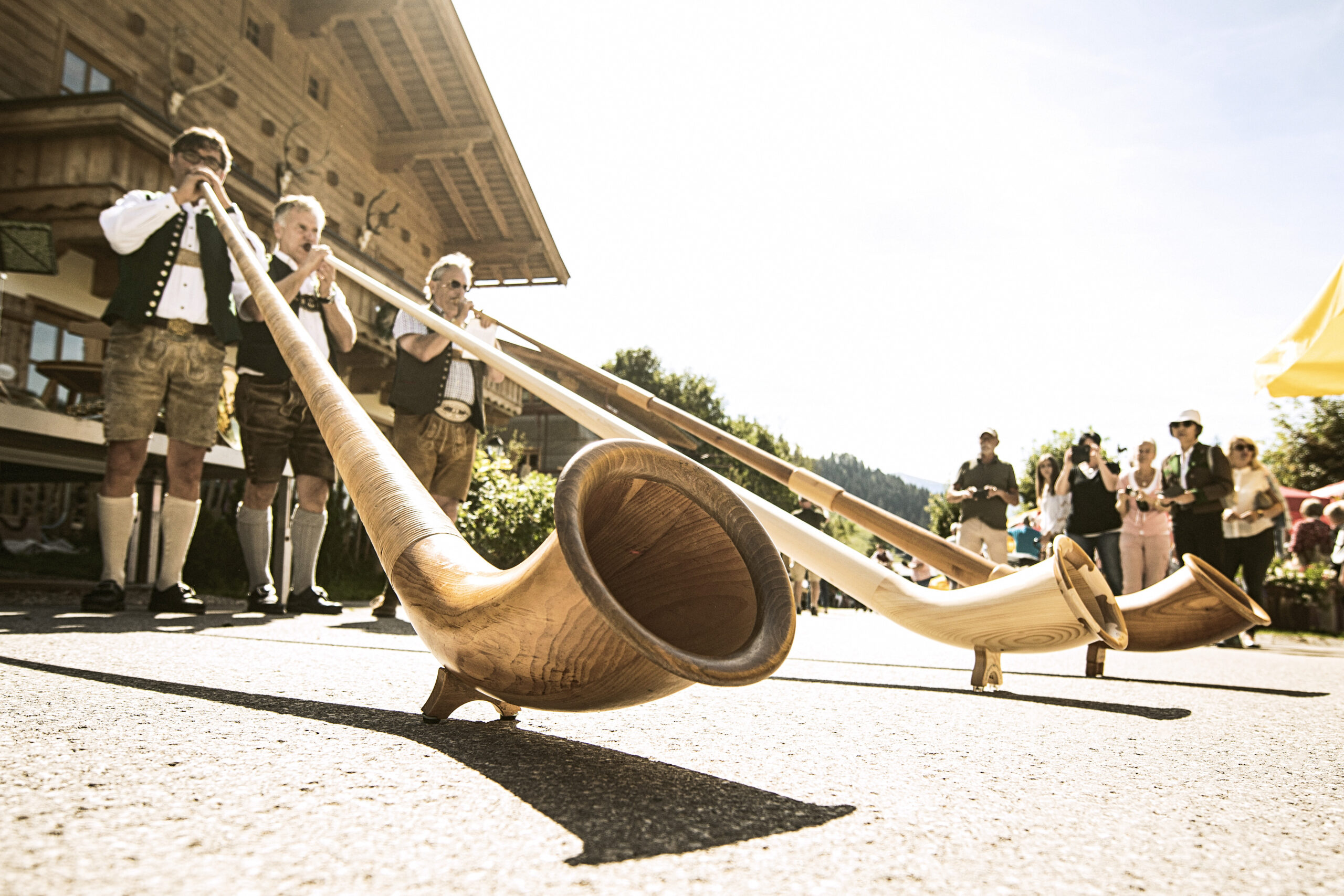Vier Personen in traditioneller österreichischer Tracht spielen bei einem Kulturfest im Freien auf langen Holzalphörnern.