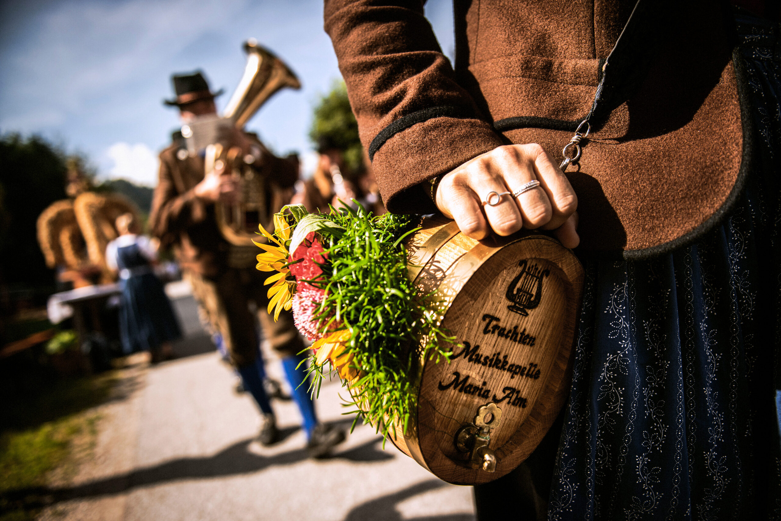 Eine Person hält bei einer Parade ein verziertes Holzfass in der Hand, im Hintergrund sind Musiker in traditioneller Tracht zu sehen.