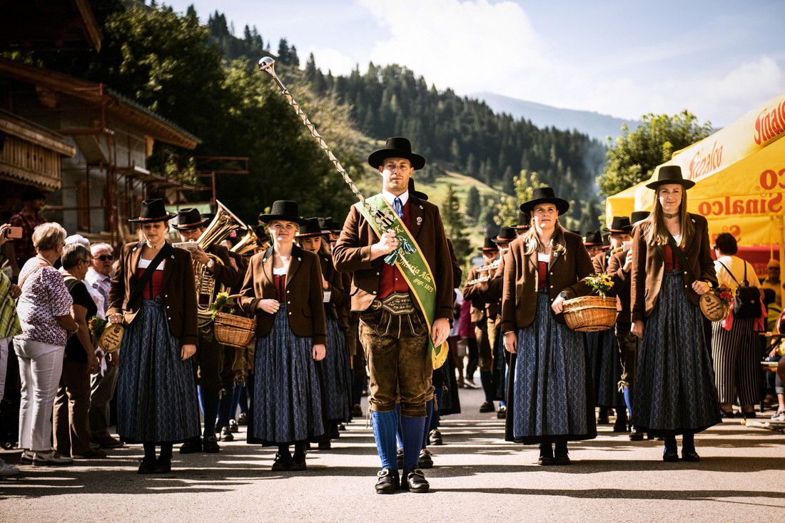 Menschen in traditioneller österreichischer Tracht ziehen in einem Umzug vorbei und tragen Körbe und Musikinstrumente.
