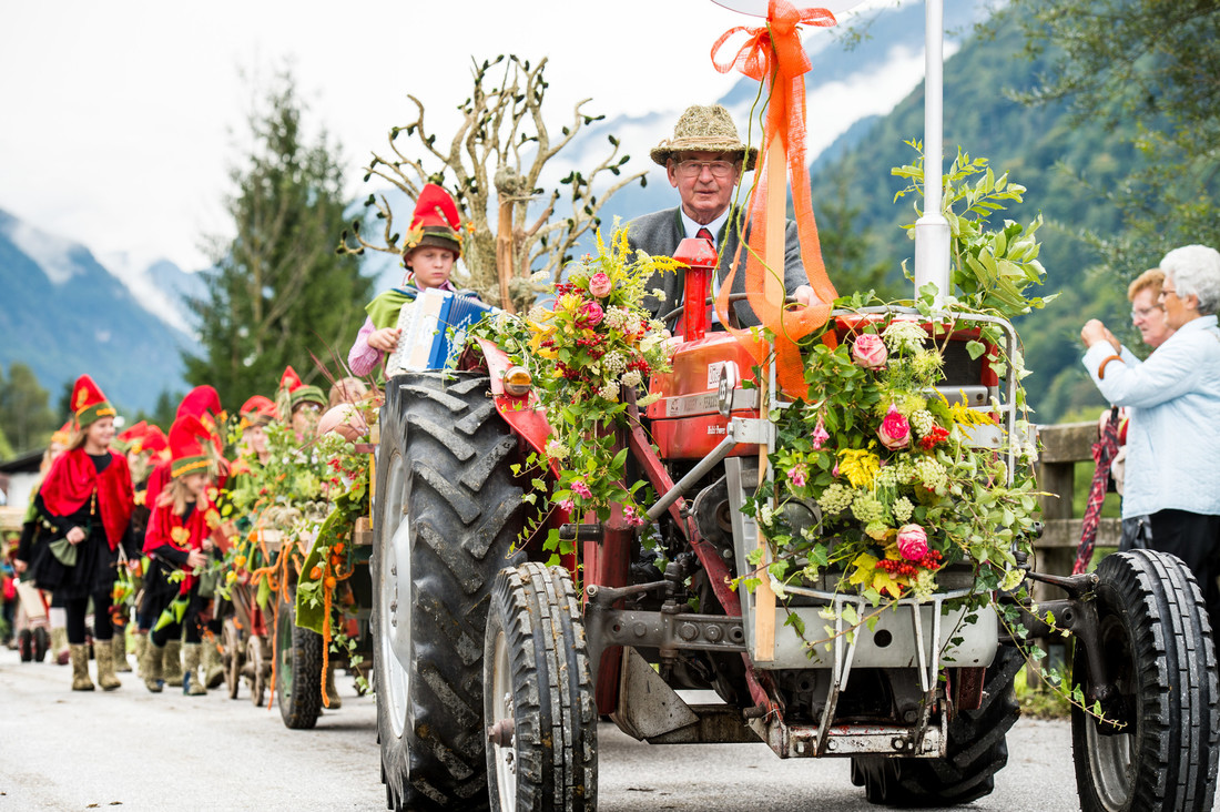 Ein Mann fährt bei einer Parade einen mit Blumen geschmückten Traktor; dahinter laufen Menschen in roten Kostümen.