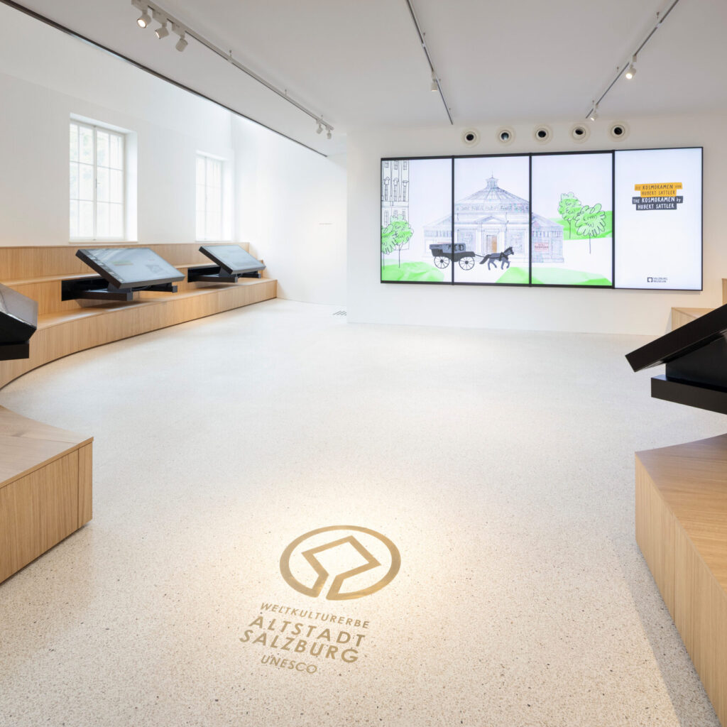 Moderner Ausstellungsraum mit interaktiven Bildschirmen, Sitzgelegenheiten aus Holz, digitaler Anzeige und dem UNESCO-Salzburg-Logo auf dem Boden.