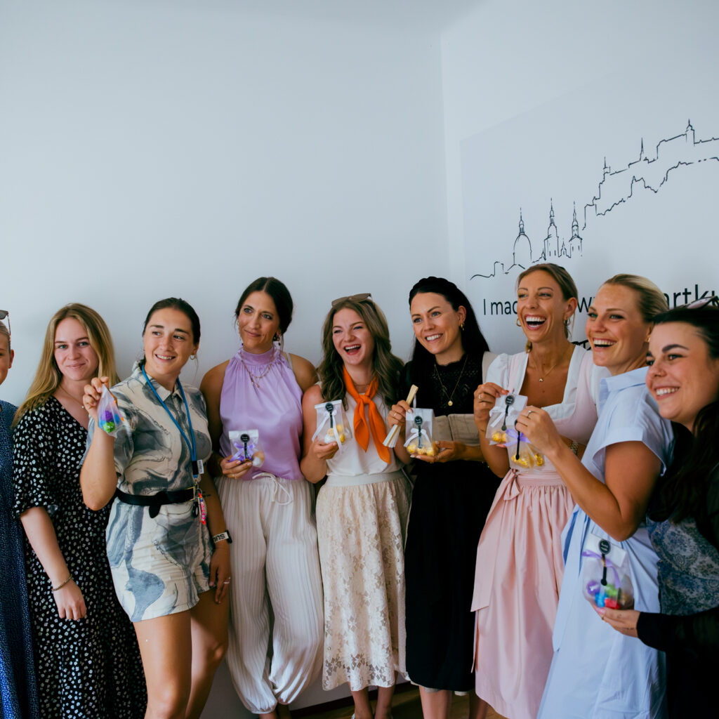 Neun Frauen in Kleidern lächeln gemeinsam in einem Raum, in den Händen Getränke und Taschen – bereit für den Social-Media-Sommer 2026.