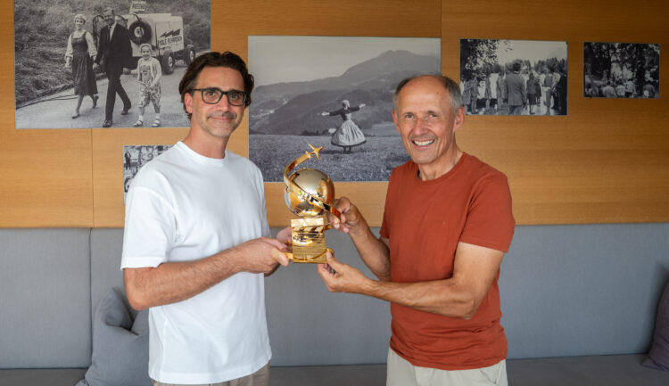 Zwei Männer, die lächeln und gemeinsam eine goldene Trophäe in den Händen halten, in einem Raum, an dessen Wänden Schwarz-Weiß-Fotos hängen.