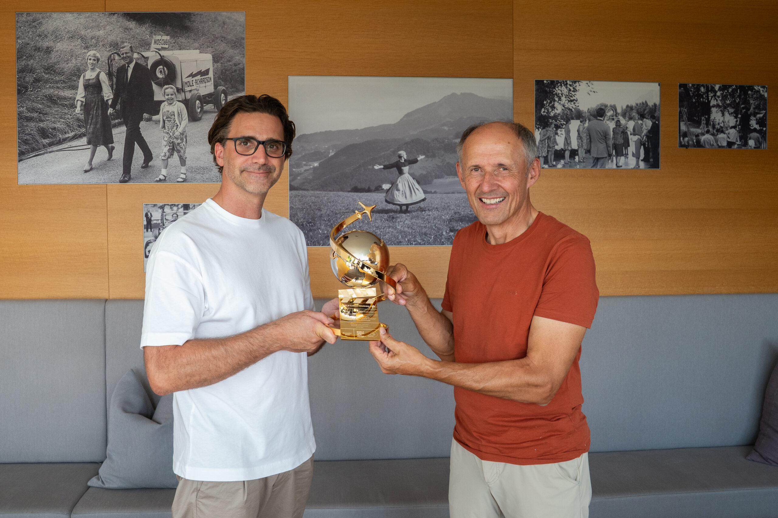 Zwei Männer, die lächeln und gemeinsam eine goldene Trophäe in den Händen halten, in einem Raum, an dessen Wänden Schwarz-Weiß-Fotos hängen.