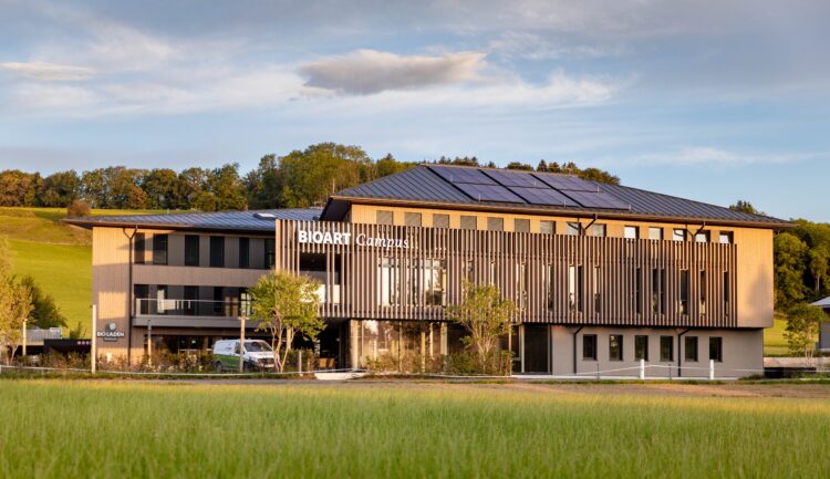 Ein modernes dreistöckiges Gebäude mit der Aufschrift „BioArt Campus“, umgeben von Solarmodulen, Bäumen und grünen Wiesen.