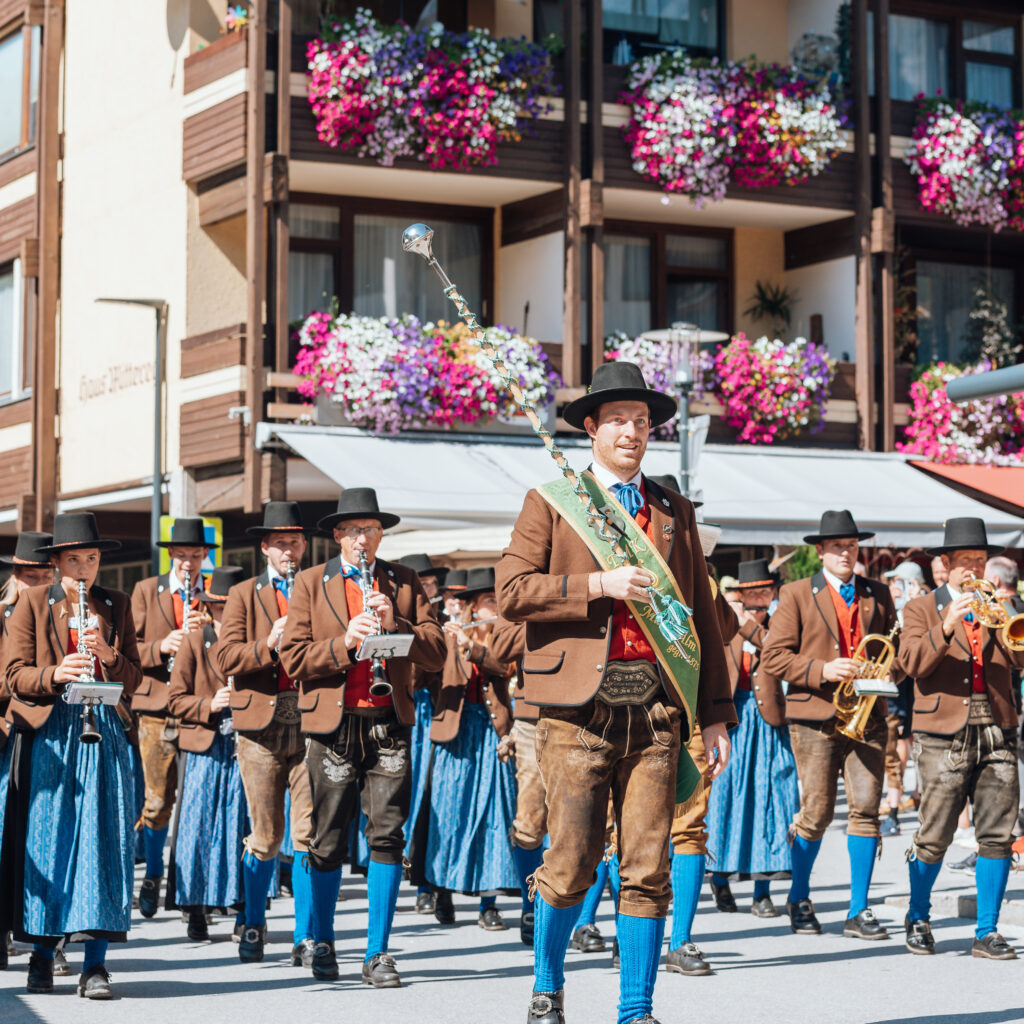 Eine Blaskapelle in traditioneller Tracht spielt auf einer sonnigen Straße, die von Gebäuden und mit Blumen geschmückten Balkonen gesäumt ist.