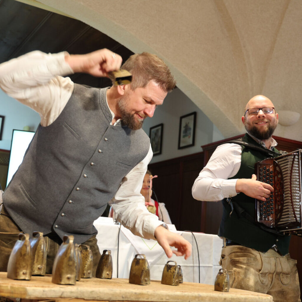 Zwei Männer in traditioneller Tracht spielen bei einer Veranstaltung in einem Saal vor Publikum Kuhglocken und Akkordeon.