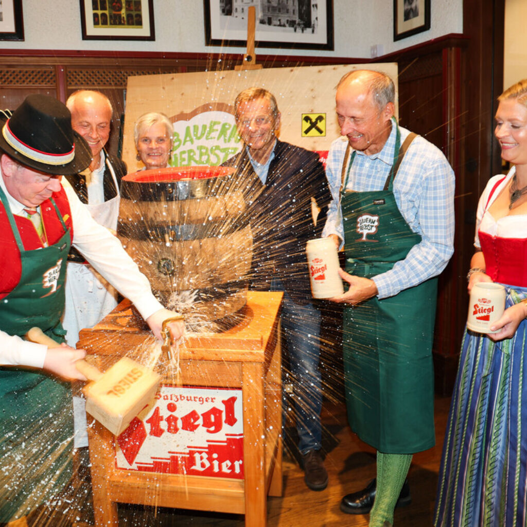 Ein Mann zapft ein Bierfass an, Schaum spritzt; andere in österreichischer Tracht lachen und halten Bierkrüge in der Nähe.