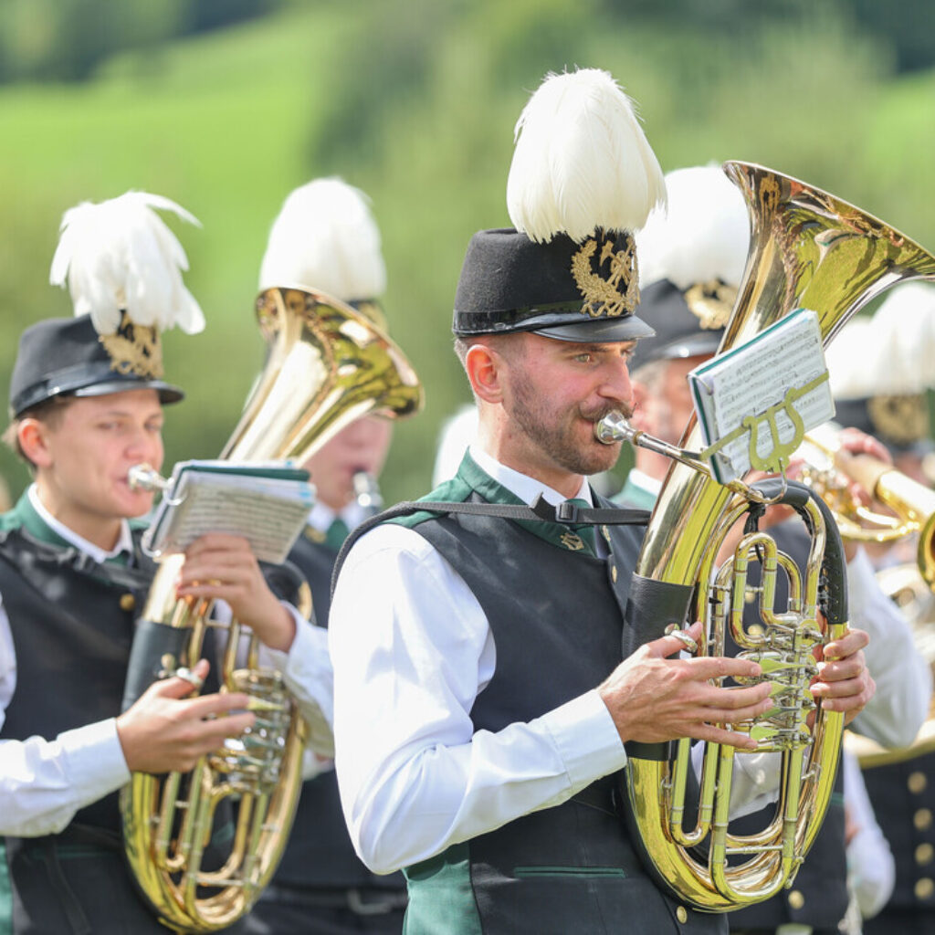 Musiker in prunkvollen Uniformen spielen im Freien in einer Blaskapelle Blechblasinstrumente, hinter ihnen erstrecken sich grüne Hügel.
