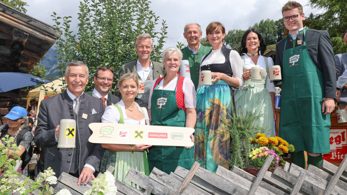 Eine Gruppe in traditioneller österreichischer Tracht steht im Freien, lächelt und hält Bierkrüge und ein Holzschild in den Händen.
