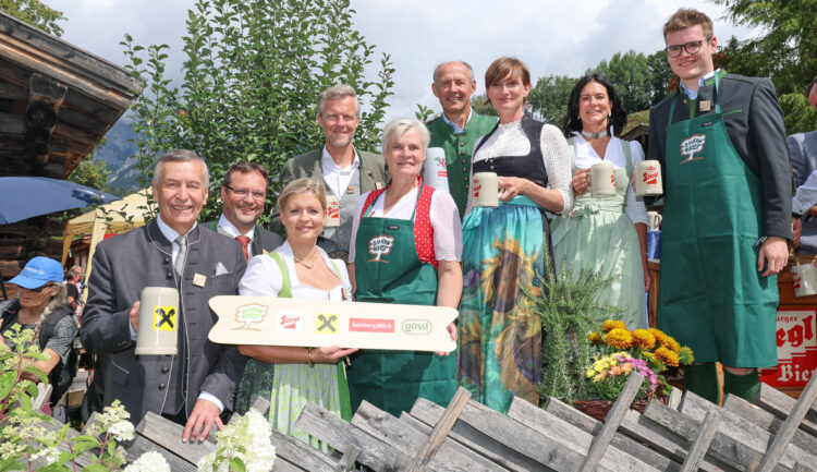 Eine Gruppe in traditioneller österreichischer Tracht steht im Freien, lächelt und hält Bierkrüge und ein Holzschild in den Händen.