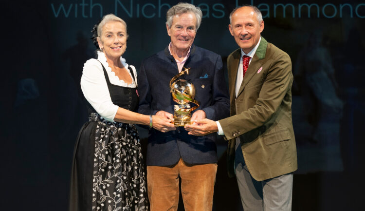 Drei Personen lächeln auf der Bühne, halten eine goldene Auszeichnung in den Händen, im Hintergrund ist ein „Sound of Music“-Schild zu sehen.