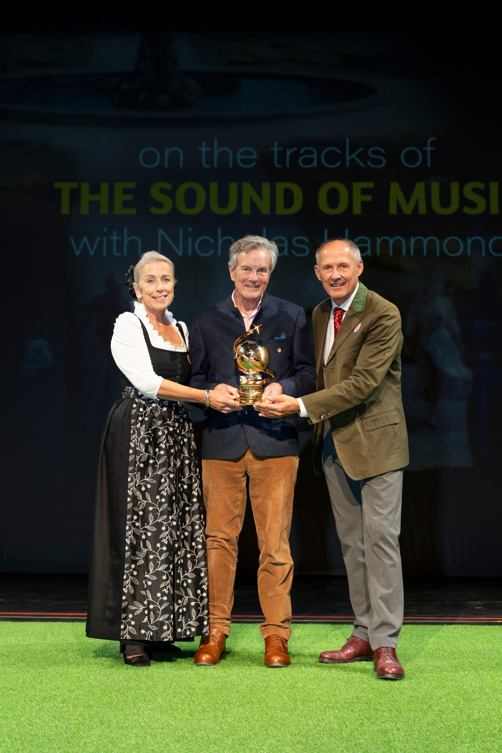 Drei Personen lächeln auf der Bühne, halten eine goldene Auszeichnung in den Händen, im Hintergrund ist ein „Sound of Music“-Schild zu sehen.