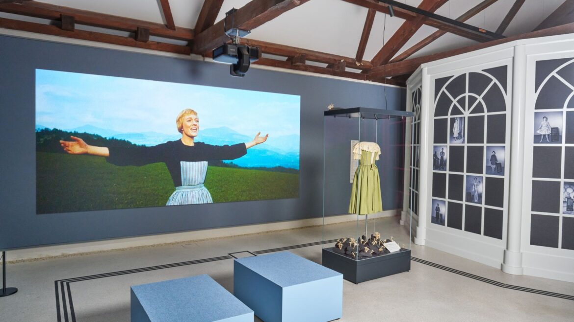 Ausstellung im „Sound of Music“-Museum in Salzburg mit einer singenden Frau, einem grünen Kleid und zwei blauen Bänken.