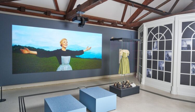 Ausstellung im „Sound of Music“-Museum in Salzburg mit einer singenden Frau, einem grünen Kleid und zwei blauen Bänken.
