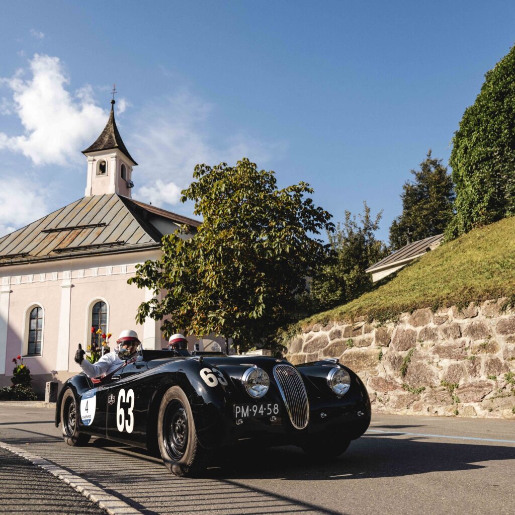 An einem sonnigen Tag fährt ein Oldtimer mit der Startnummer 63 an einer Kirche vorbei; darin sitzen zwei Personen.