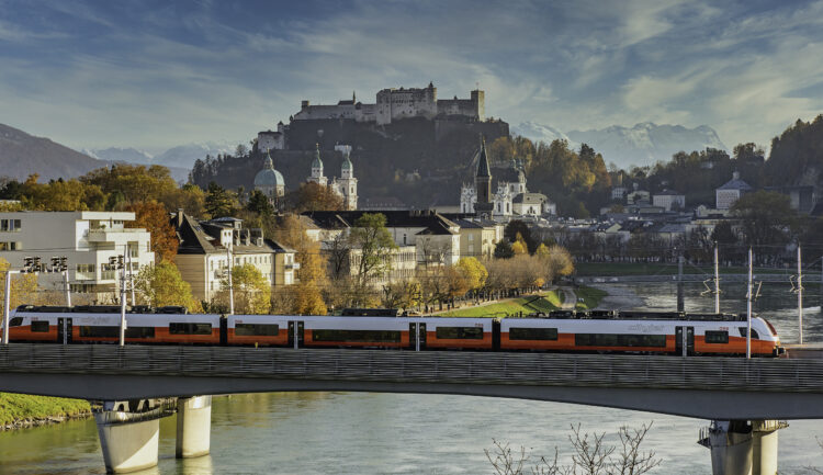 Ein roter Zug überquert eine Brücke in Salzburg, Österreich – die perfekte Kulisse für eine Umfrage zum „Guest Mobility Ticket“.