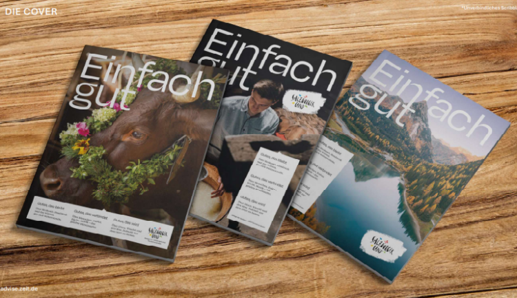 Drei Ausgaben der Zeitschrift „Einfach gut“ auf einem Holztisch, auf deren Titelseiten eine Kuh, ein Koch und ein See abgebildet sind.