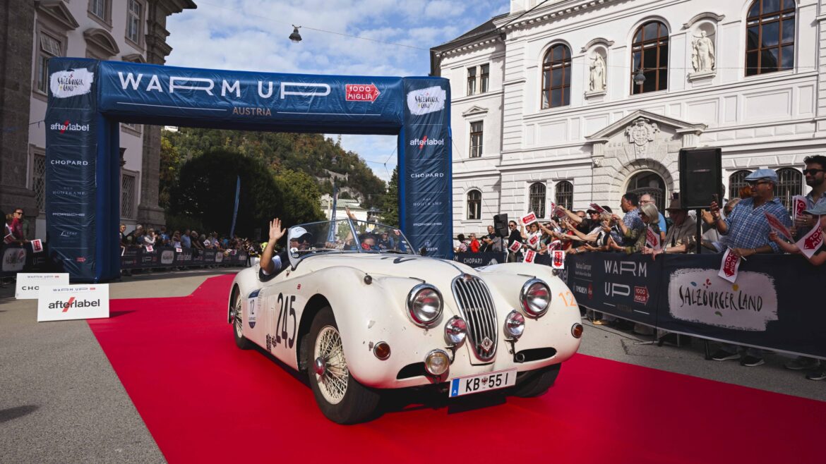 Ein klassischer weißer Wagen fährt bei einer Rallye über einen roten Teppich, im Hintergrund jubelt und winkt die Menge.