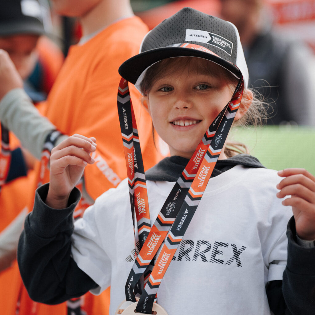 Ein lächelndes Kind mit einer Mütze hält eine INFINITE TRAILS-Medaille bei der Outdoor-Veranstaltung „Event-Rückblick September 2026“ in den Händen.