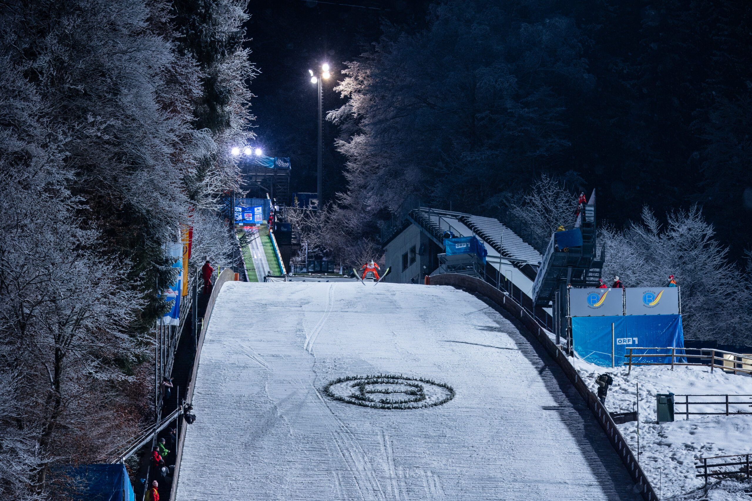Ein Skispringer in der Luft, der nachts einen beleuchteten, schneebedeckten Hang hinunterfliegt.