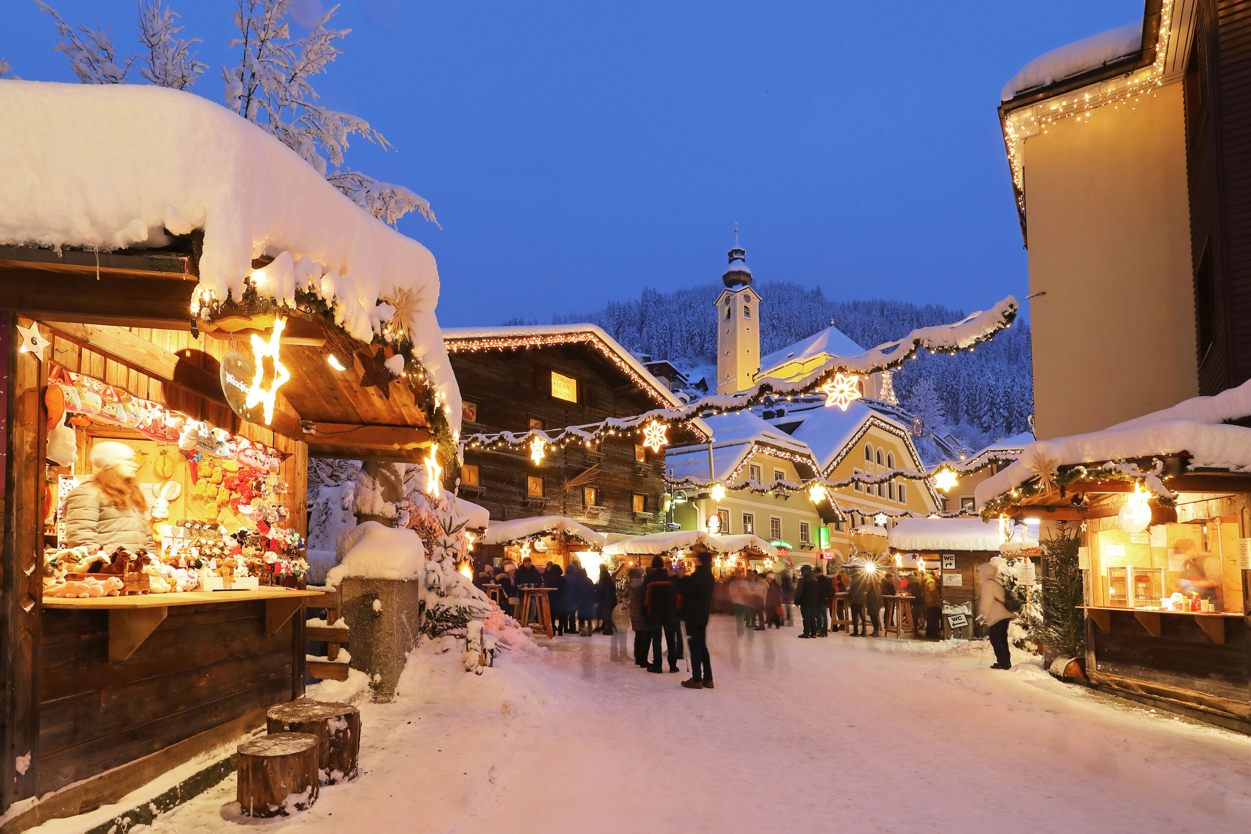 Menschen, die in der Dämmerung einen österreichischen Weihnachtsmarkt im Freien in einem verschneiten Dorf besuchen.