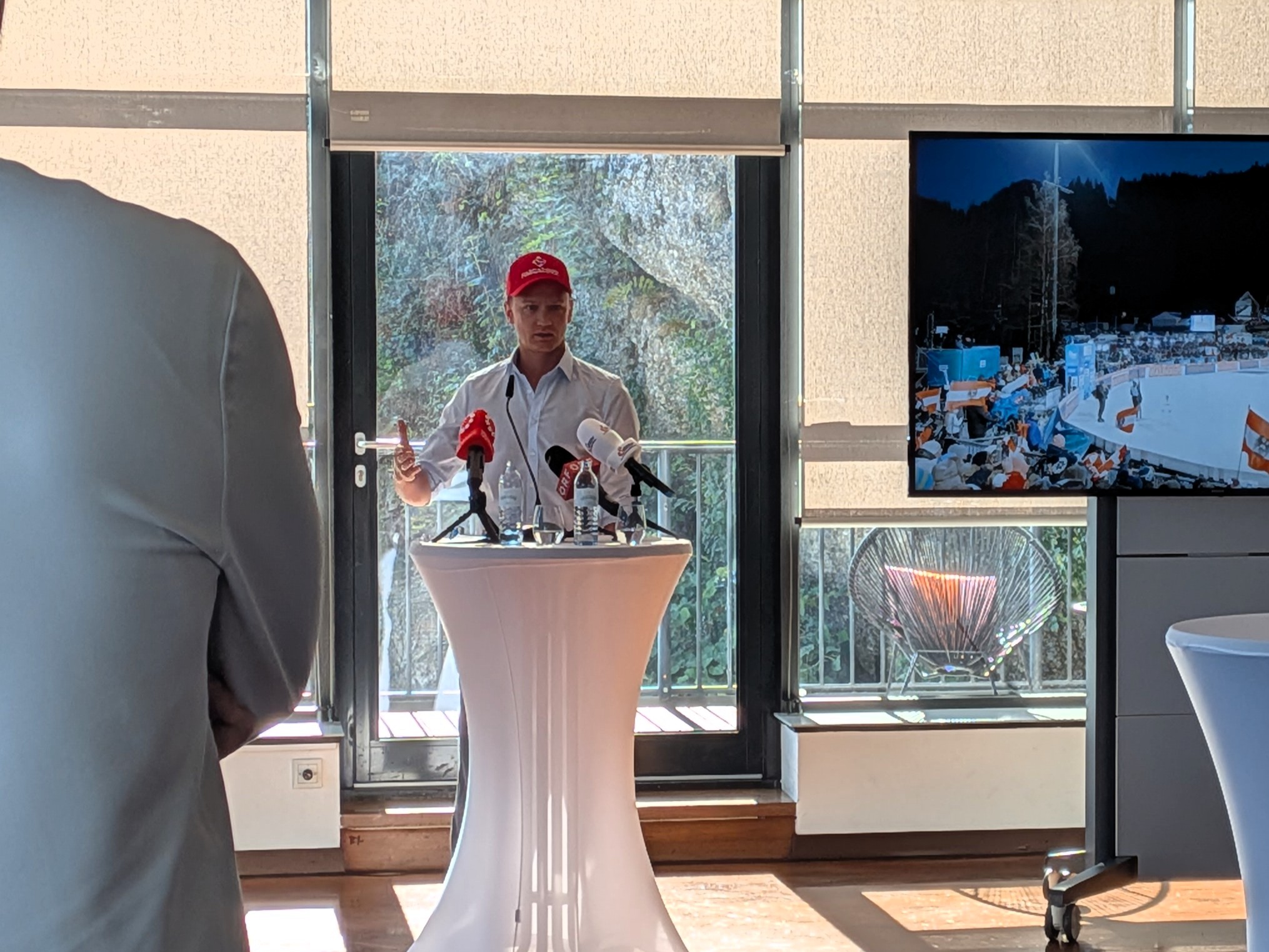 Ein Mann mit einer roten Mütze steht an einem Rednerpult mit Mikrofonen und spricht vor einer Leinwand, auf der ein verschneites Stadion zu sehen ist.