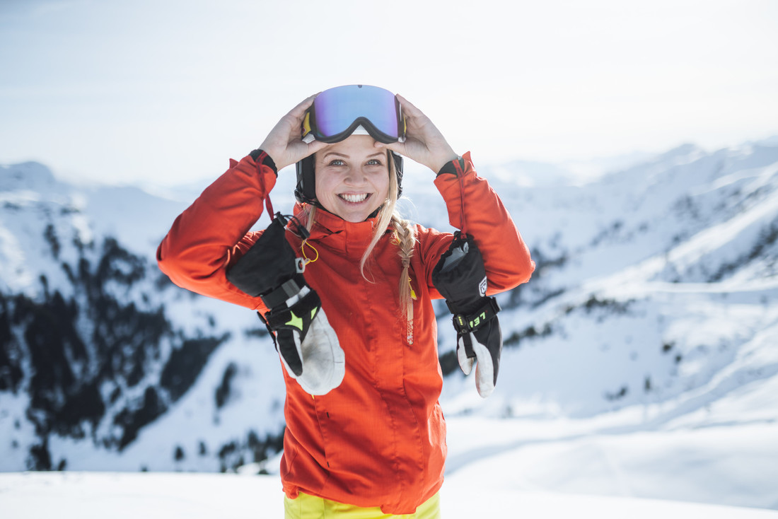 Ein lächelnder Skifahrer in einer roten Jacke und mit einer Skibrille, der auf einem verschneiten Berg steht.