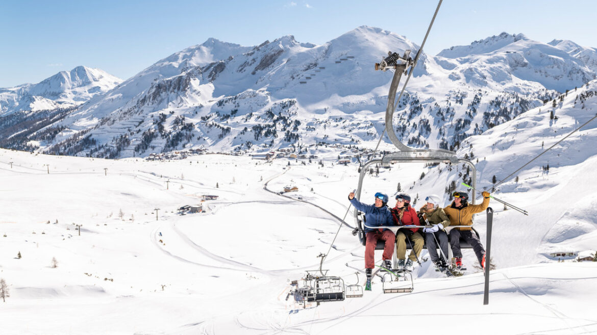 Vier Skifahrer sitzen auf einem Sessellift und machen ein Selfie vor der Kulisse der schneebedeckten österreichischen Berge.