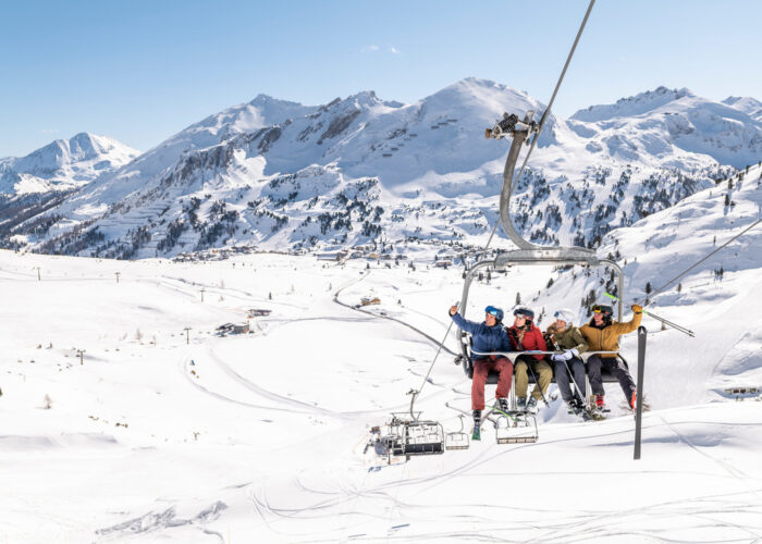 Vier Skifahrer sitzen auf einem Sessellift und machen ein Selfie vor der Kulisse der schneebedeckten österreichischen Berge.