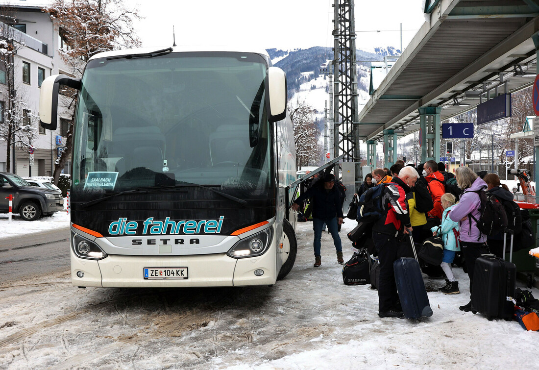 Reisende mit Gepäck warten an einer verschneiten Bushaltestelle in einem österreichischen Bergort darauf, in einen Reisebus einzusteigen.