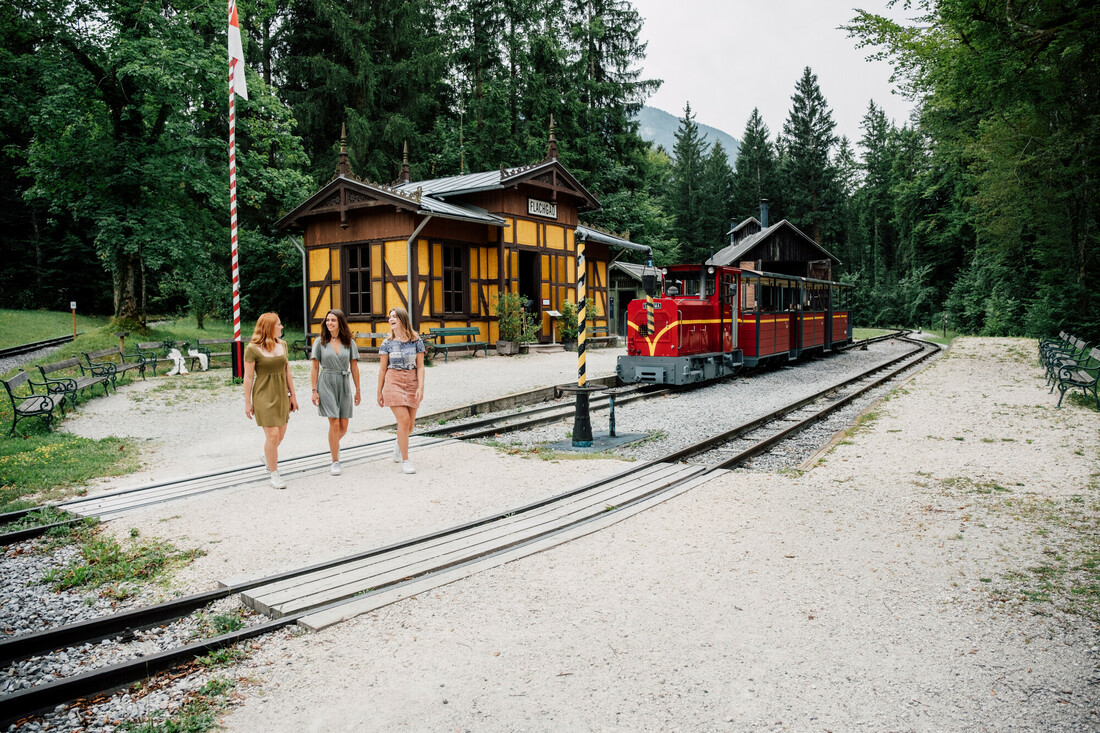 Drei Frauen gehen an einem roten Zug vorbei, der an einem kleinen Bahnhof im Wald hält.