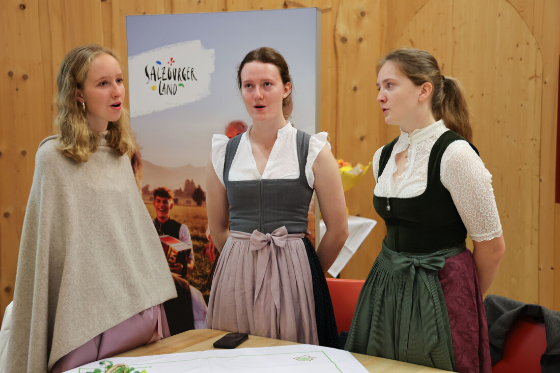 Drei Frauen in traditioneller österreichischer Tracht singen gemeinsam vor einem Werbebanner.