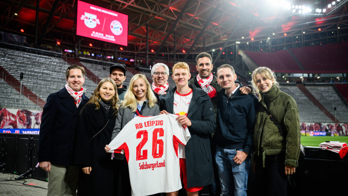 Eine Gruppe von Menschen steht auf einem Fußballplatz und hält ein weißes Trikot mit der Nummer 26 von RB Leipzig auf dem Rücken hoch.