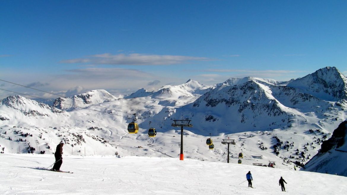 Skifahrer auf einem verschneiten Berghang mit Skiliften und schneebedeckten Gipfeln im Hintergrund unter blauem Himmel.