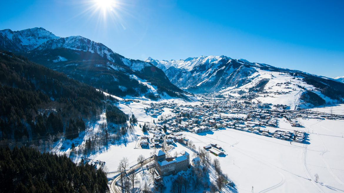 Ein sonniges, verschneites Alpendorf mit Bergen im Hintergrund und einem Wald im Vordergrund.