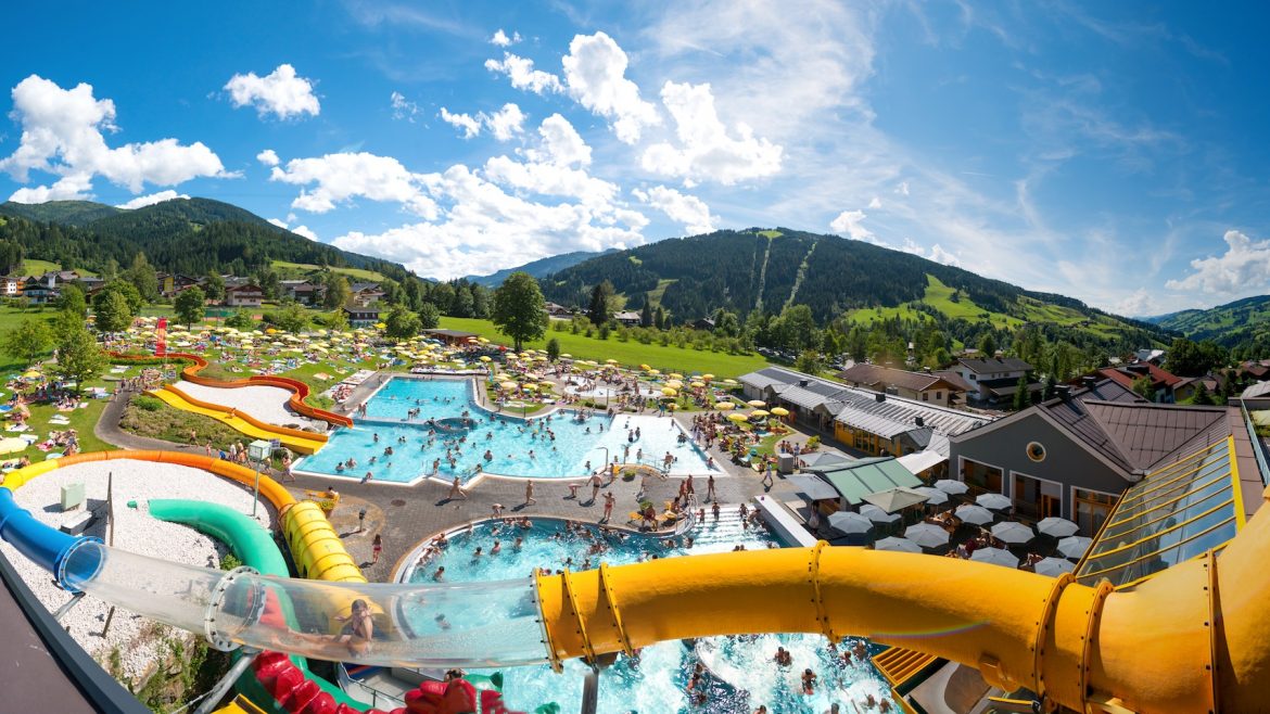Ein großer Wasserpark im Freien mit Becken, Wasserrutschen und Menschen, eingebettet in eine malerische grüne Berglandschaft.