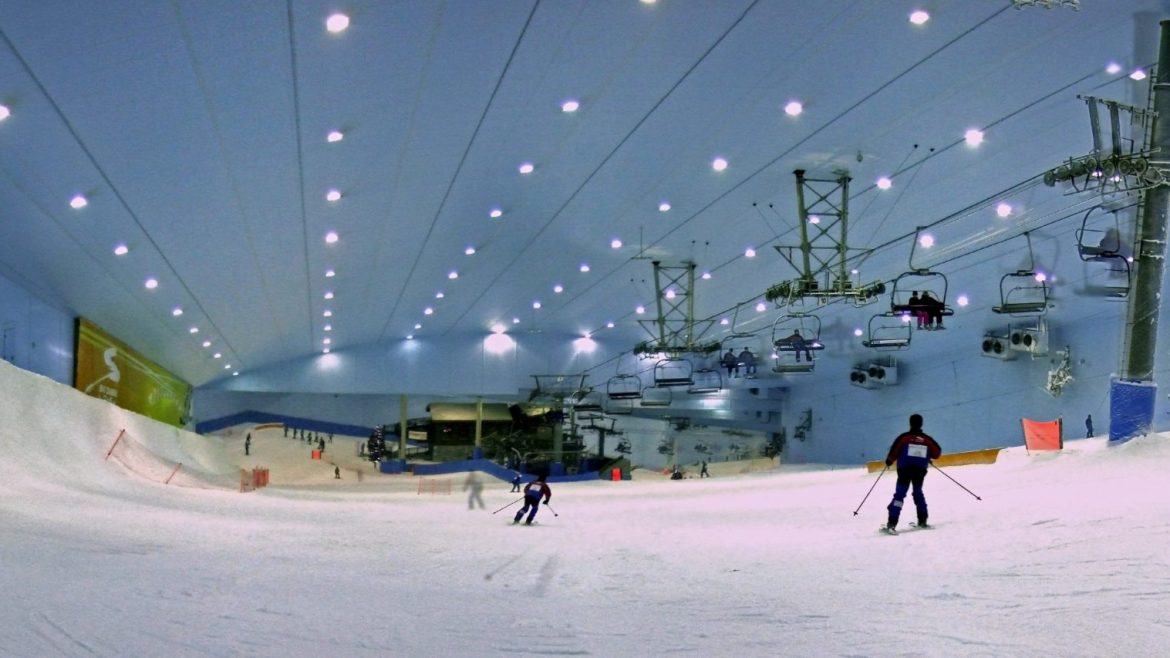 Indoor-Skipiste mit Skifahrern auf dem Schnee und Sesselliften über dem Kopf unter heller Deckenbeleuchtung.